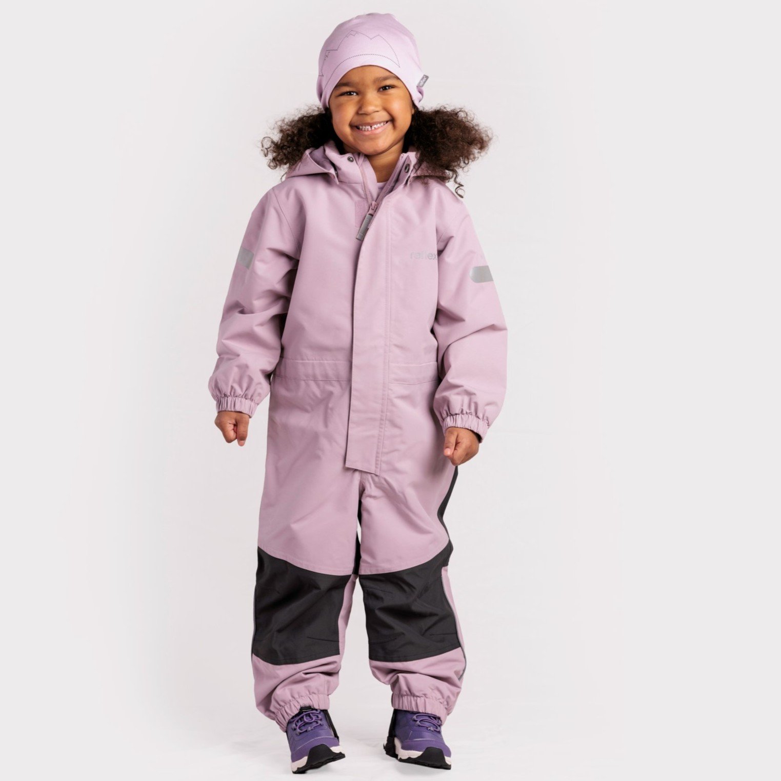 Barn har på seg rosa hetteoverall med svarte benpaneler og refleksdetaljer, rosa mønstret lue og lilla joggesko, forfra.