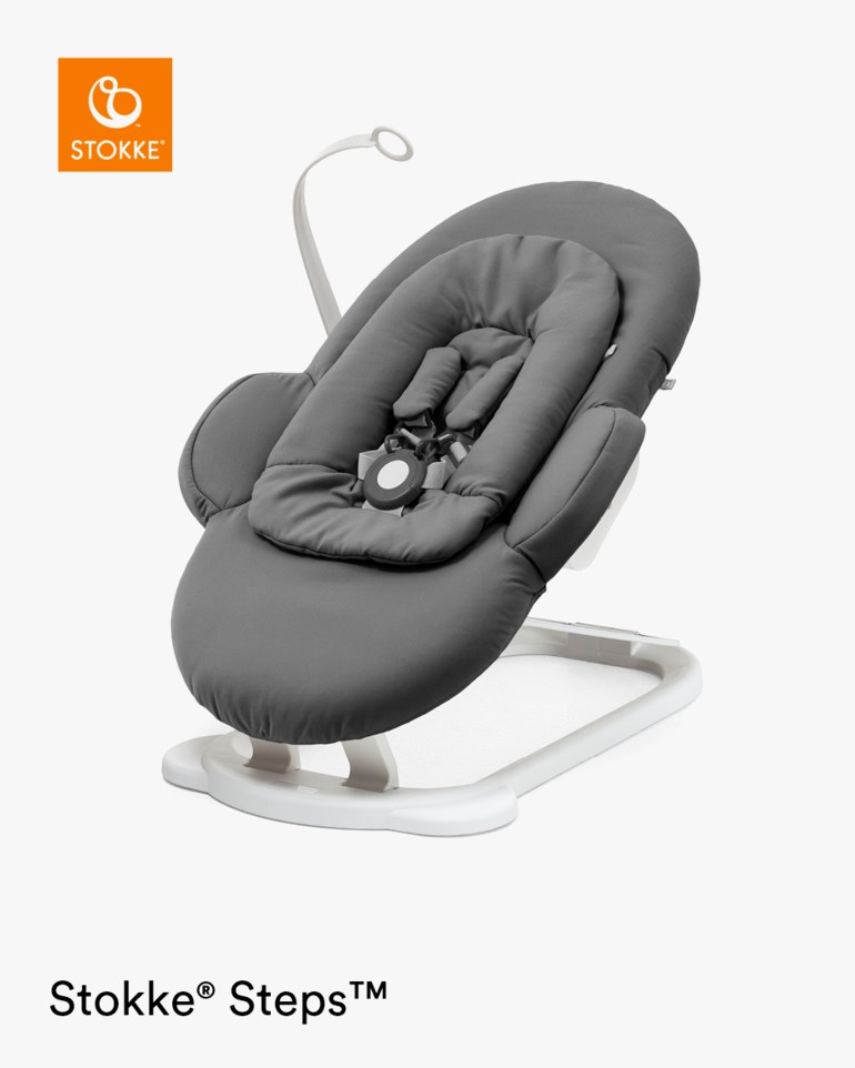 En mørkegrå vippestol med hvit ramme, polstret innlegg, 5-punktssele og en leketøybue.