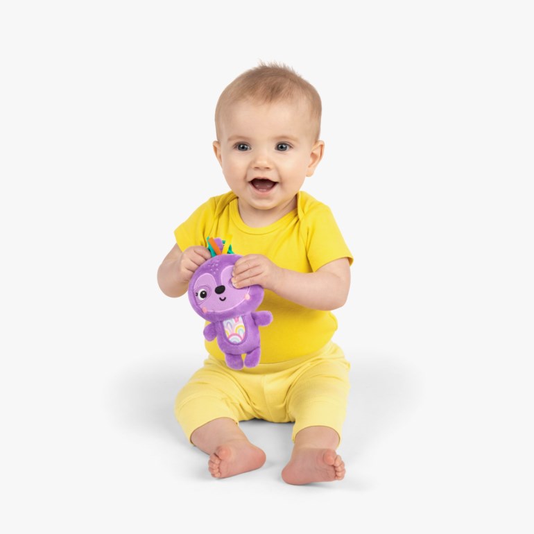 Baby holder et lilla plysjdyr med regnbuedetaljer, iført en gul kortermet body og gule bukser.