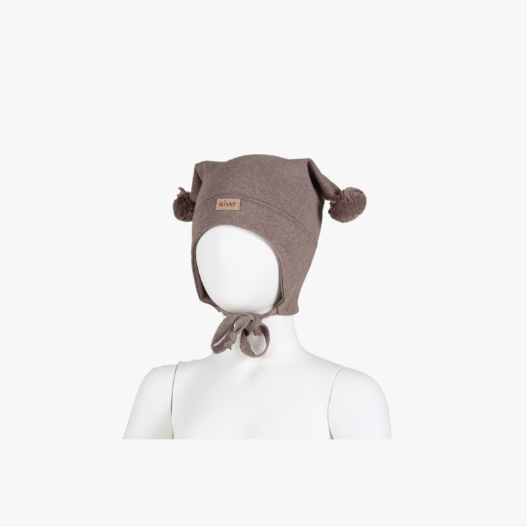 Brun strikket ørelappslue for barn med to dusker og knytebånd, båret av en barnedukke.