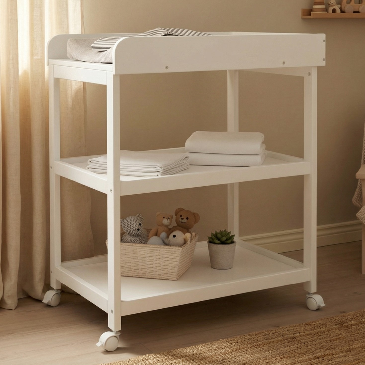 Hvitt stellebord på hjul med tre hyller, stellematte, brettede tepper, kosedyr og en plante.