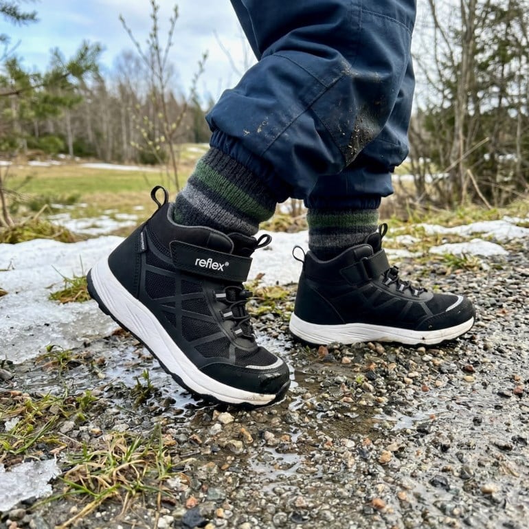 Barnets svarte tursko med hvit såle og borrelåsfeste, brukt med stripete sokker og blå bukser.