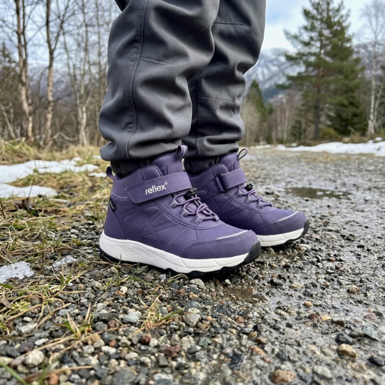 Barn bruker lilla høye utendørssko med borrelås, hvite såler og grå bukser utendørs.