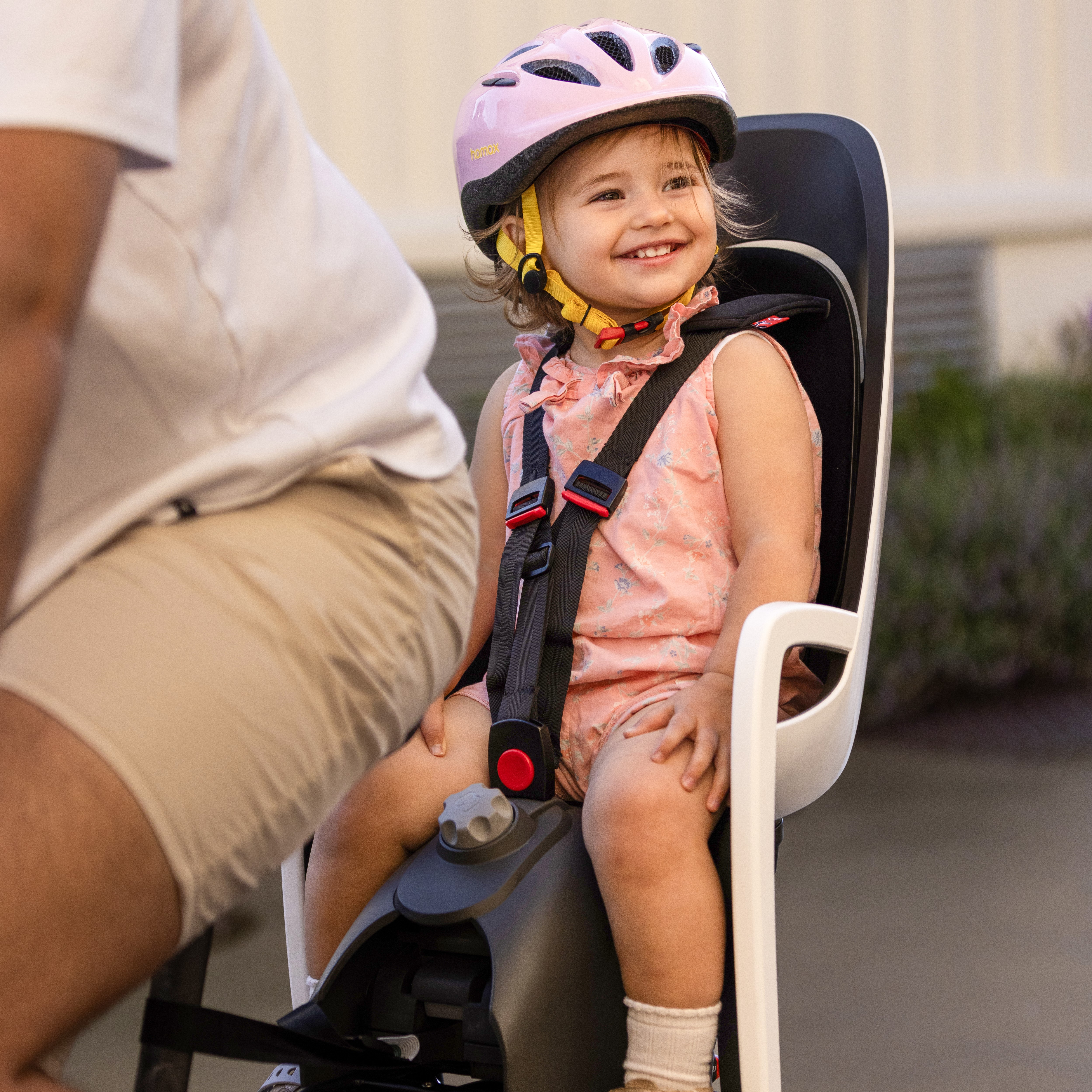 Barn i svart-hvit sykkelsete med svarte stropper, bærer rosa hjelm. Voksen har lysebrune shorts.