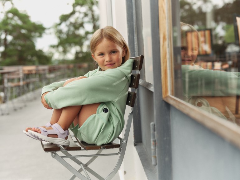 Barn bærer lysegrønn genser med matchende shorts og lys lilla borrelåssandaler, sittende.