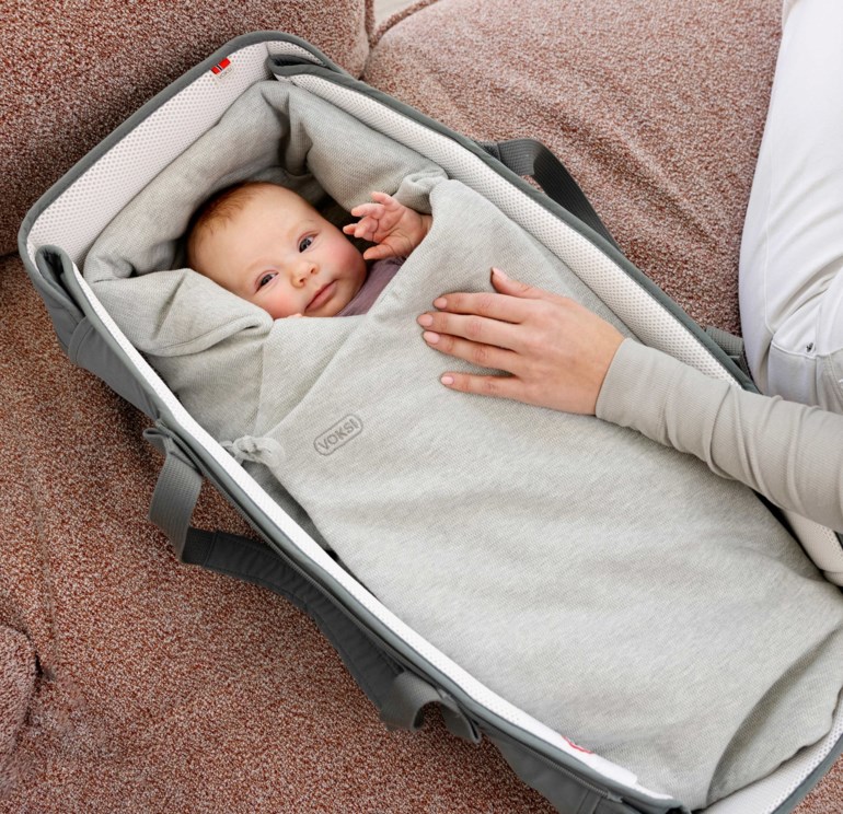 Myk grå babylift med en baby inni; en kvinnes hånd hviler på den.