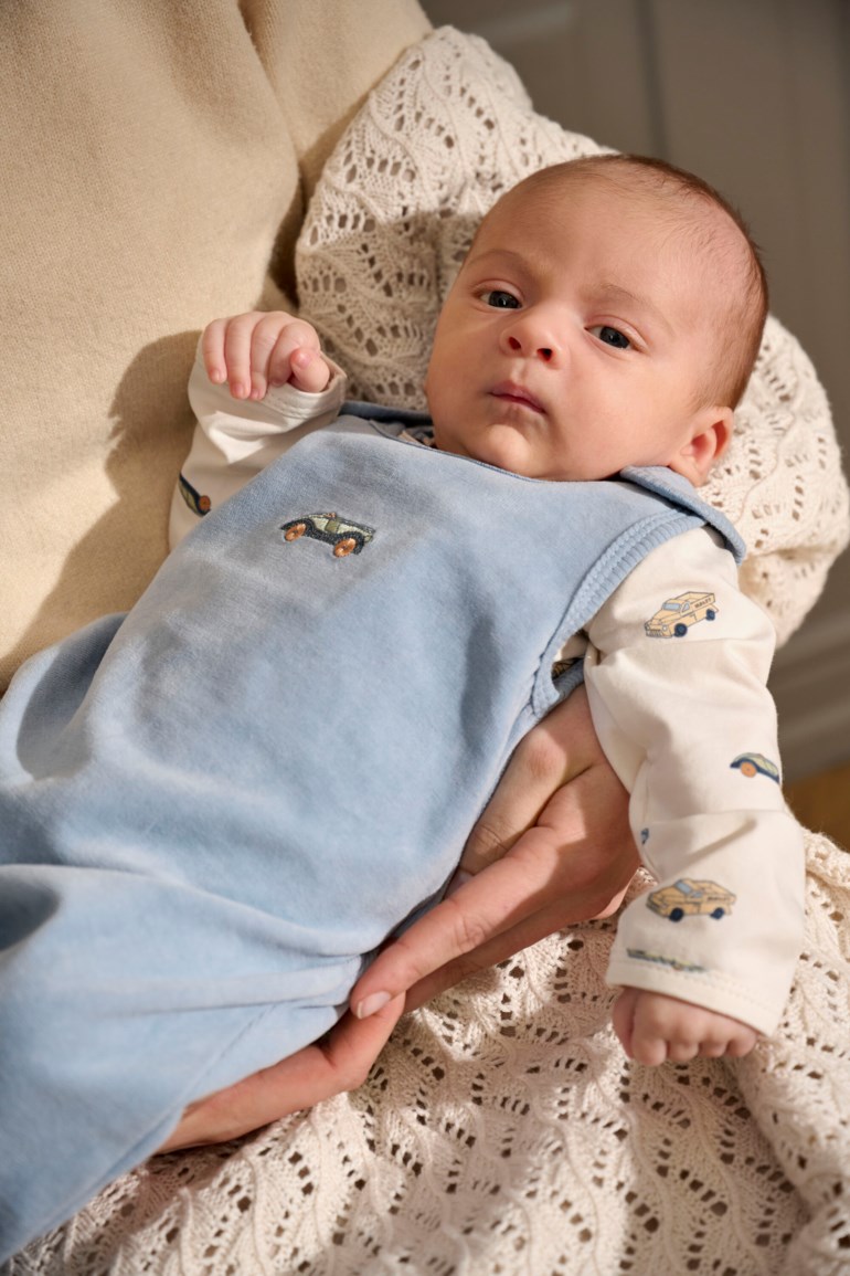 Babyen har på seg en lyseblå ermeløs plysjdress med bilbroderi over en kremfarget mønstret langermet topp.