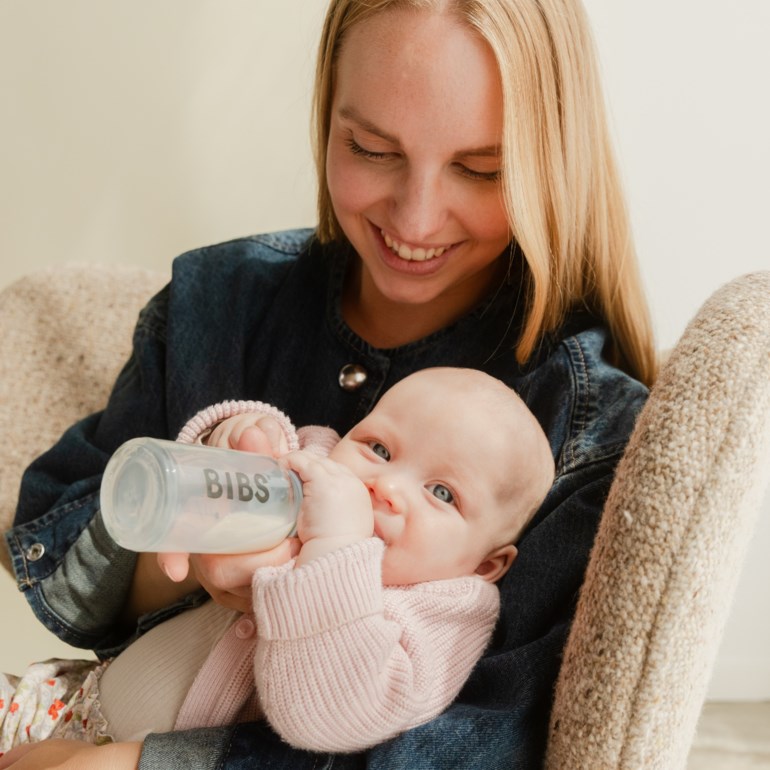 Gjennomsiktig tåteflaske med hvit væske holdt av en baby i rosa strikket cardigan og en kvinne i dongerijakke.