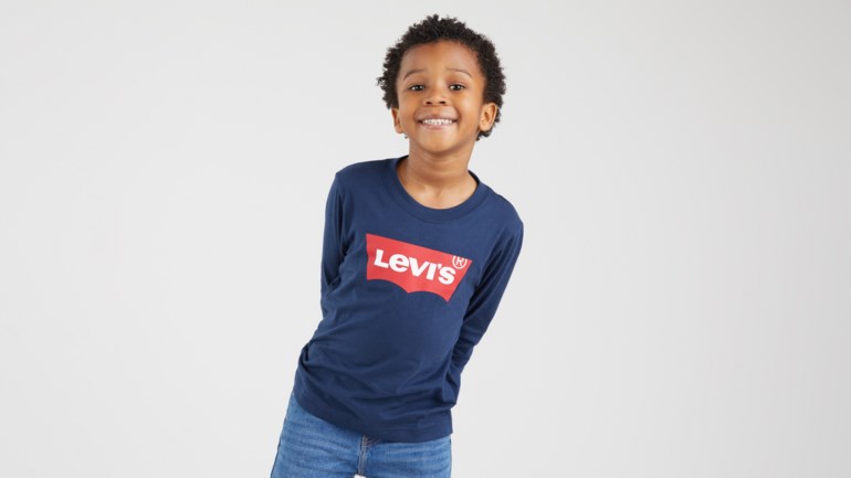 Et barn har på seg en mørkeblå langermet t-skjorte med rød logo på brystet og lyseblå jeans, forfra.