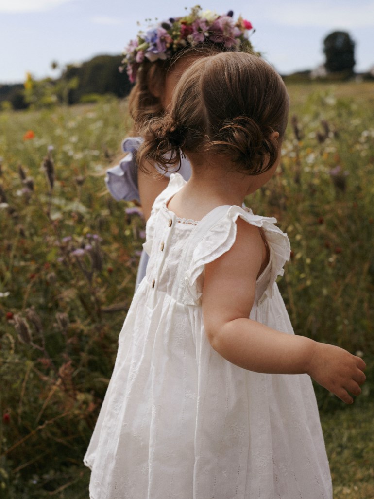 Barnes hvitbroderte kjole med volangermer og knapper bak; et annet barn bærer en blomsterkrans.