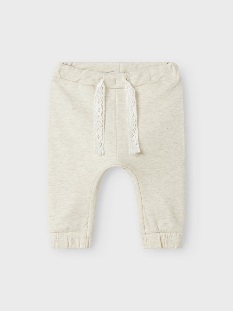 Frontvisning av lys beige babybukse med elastisk midje, dekorativ hvit blondesnor og elastiske mansjetter.