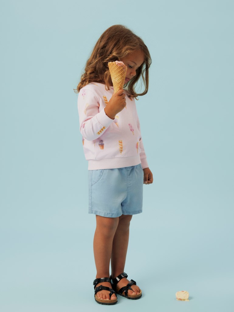 Rosa langermet genser med ismønster, lyseblå shorts og sorte sandaler på barn som holder en is.
