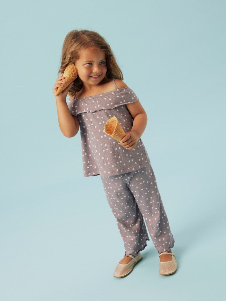 Barn i grå prikkete off-shoulder topp og matchende bukser, holder to iskremkjeks, med lysebeige flate sko.