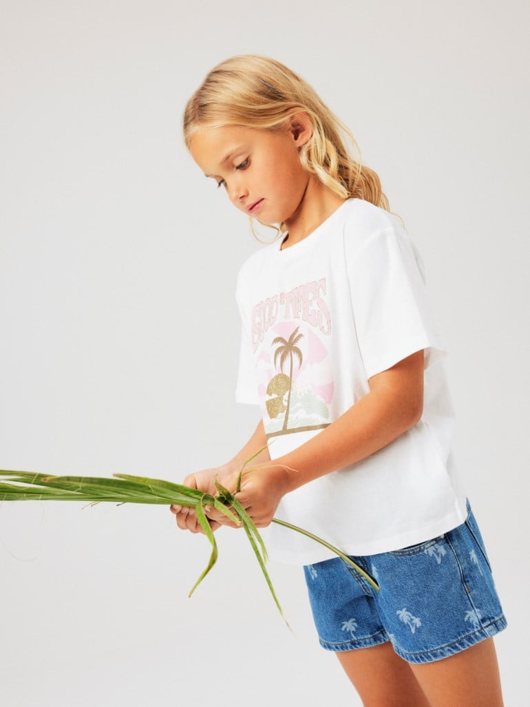 Barn har på seg hvit 'GOOD VIBES' grafisk t-skjorte og blå denimshorts med palmemønster, holder grønne blader.