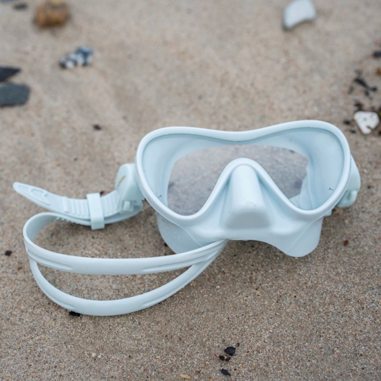 En lyseblå dykkermaske med klare linser og en silikonrem ligger på sand.