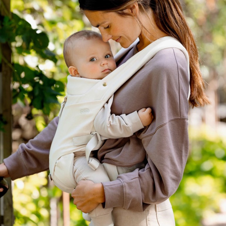 Beige bæresele båret av kvinne som bærer baby, over brun genser og beige bukser.