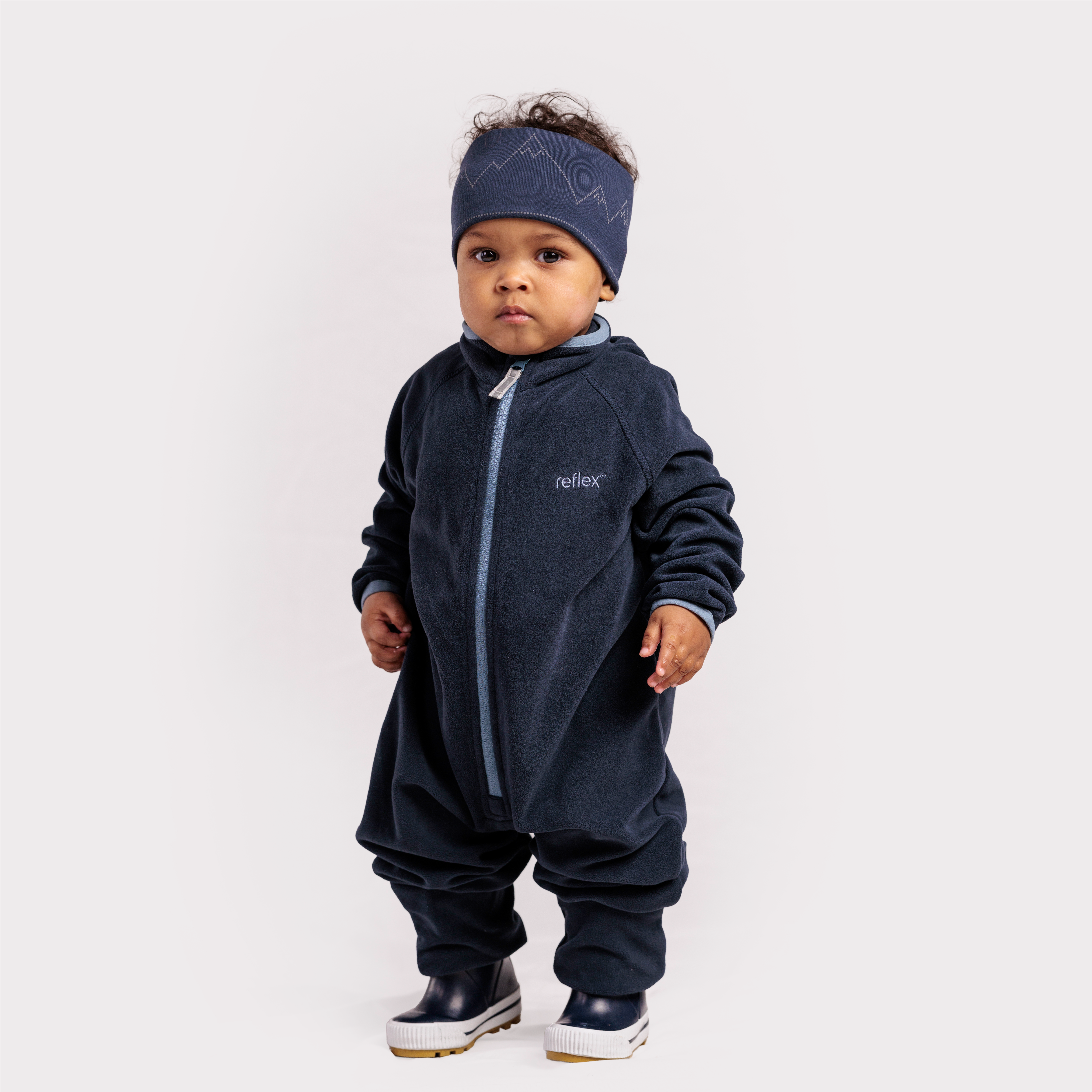 Baby har på seg en mørkeblå fleeceoverall med lyseblå glidelås, et matchende pannebånd med fjellmønster og mørkeblå støvler.