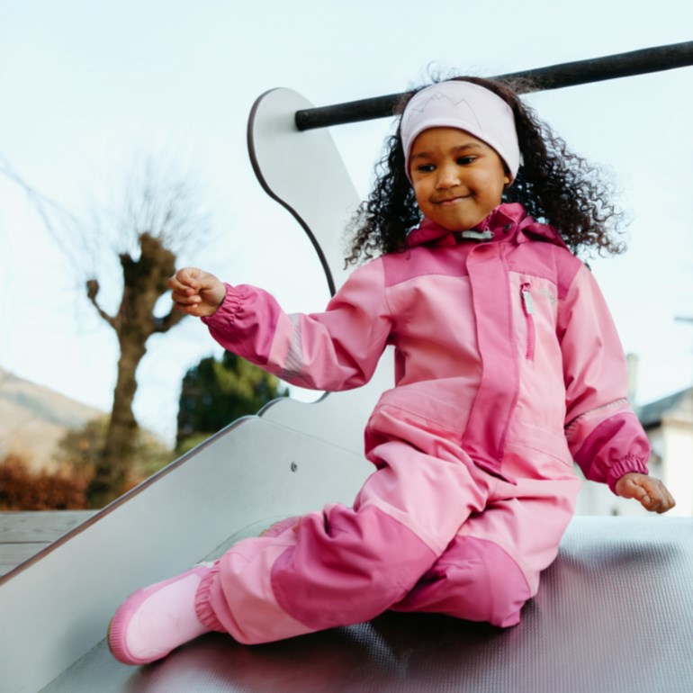 Barn i rosa tofarget snøoverall og hvitt pannebånd med fjelltrykk, sittende på en lekeplassrutsjebane.