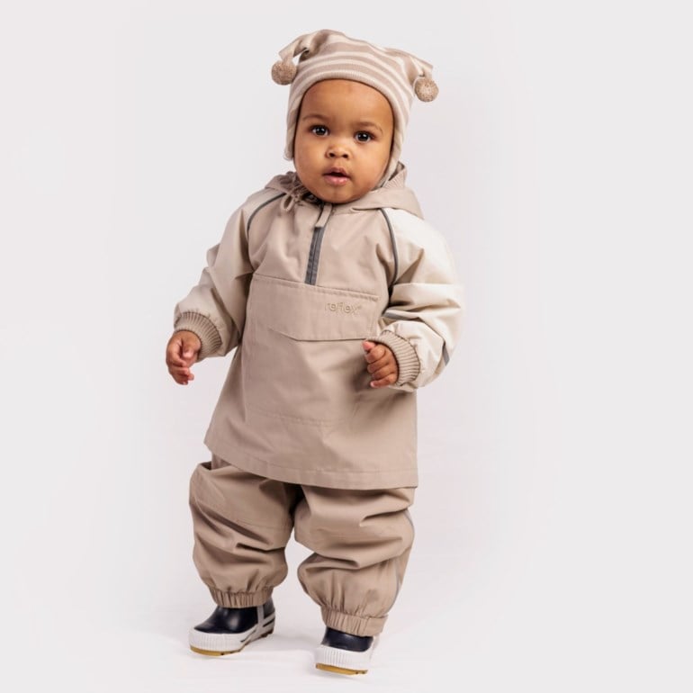 Baby har på seg en beige hettejakke, matchende bukse, en stripet lue med dusker og svarte støvler.