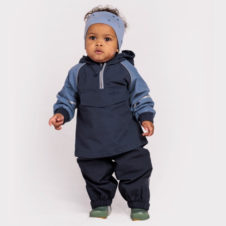 Baby bærer mørkeblå og lyseblå anorakkjakke, matchende bukser, mønstret blått pannebånd og grønne støvler.