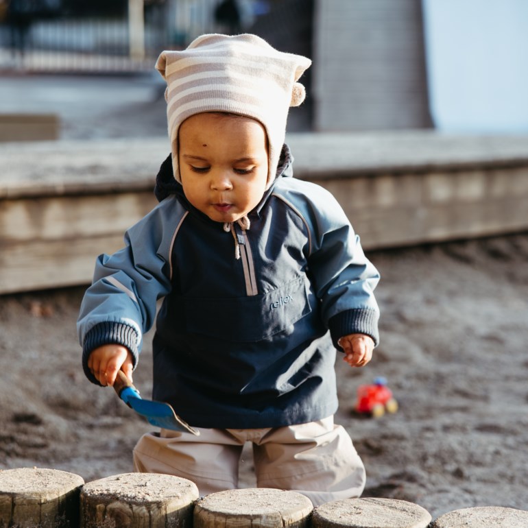Barn har på seg en blå og marineblå anorakkjakke med halvglidelås, stripete lue og lysebeige bukser, holder en blå spade.