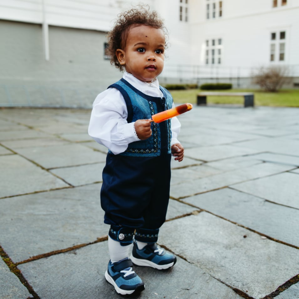 Baby i blå brodert vest, bukser, hvit skjorte, hvite sokker og blå joggesko, holder en ispinne.