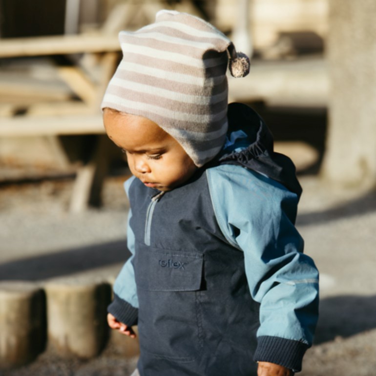 Baby bærer en mørkeblå og lyseblå jakke med frontlomme og en stripete beige lue med dusk.