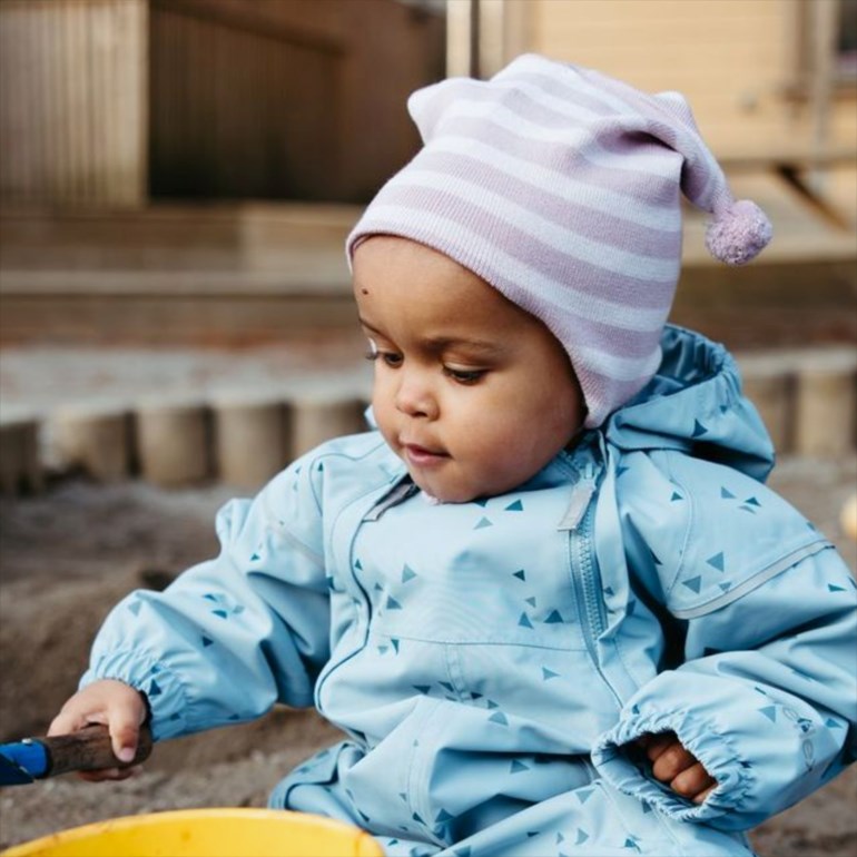 Baby i lyseblå trekantmønstret vanntett overall, rosa/hvit stripete lue, holder pinne i sand.