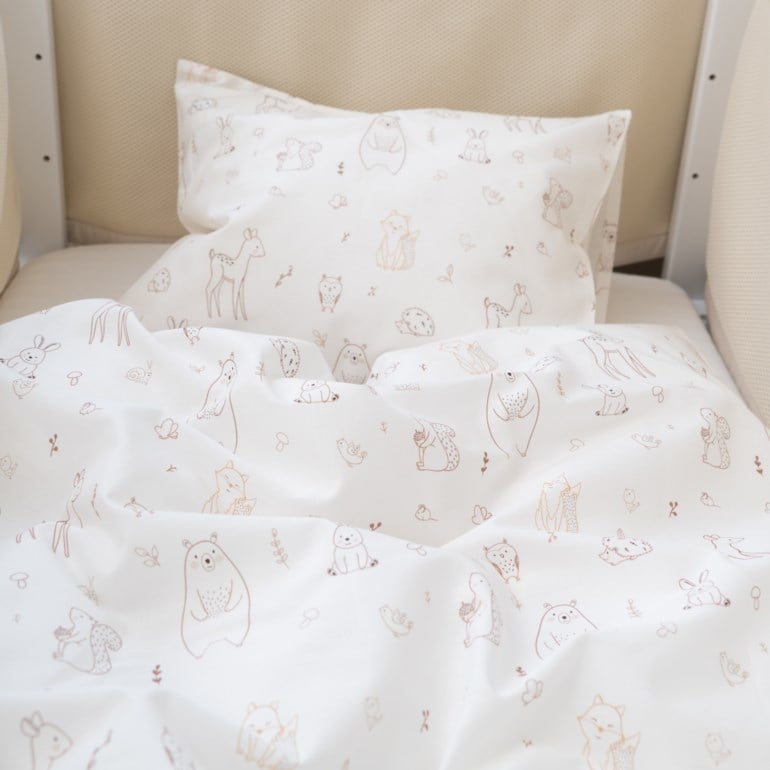 Hvitt baby sengetøy med lysbrunt skogsdyrmønster og beige netting sengekantbeskytter.