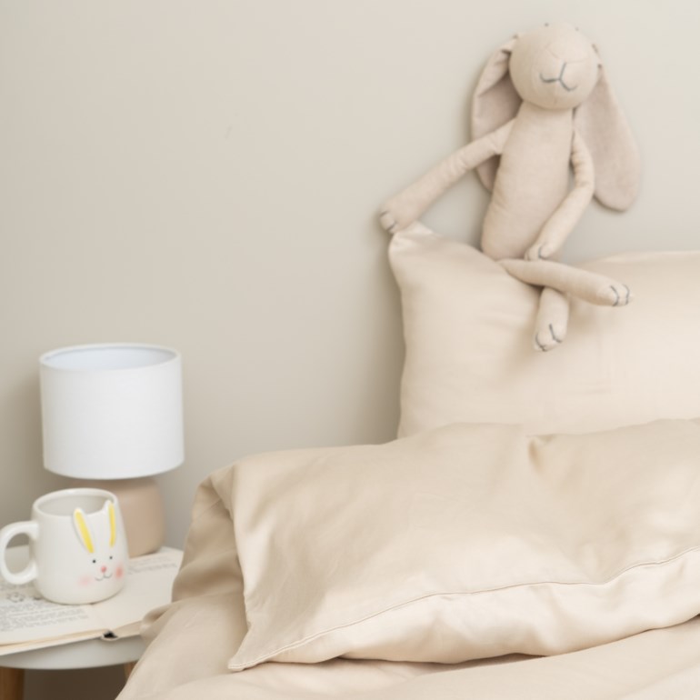 Beige sengetøy, inkludert et dynetrekk og putetrekk, med en beige kosedyrkanin, hvit lampe og kaninkopp.