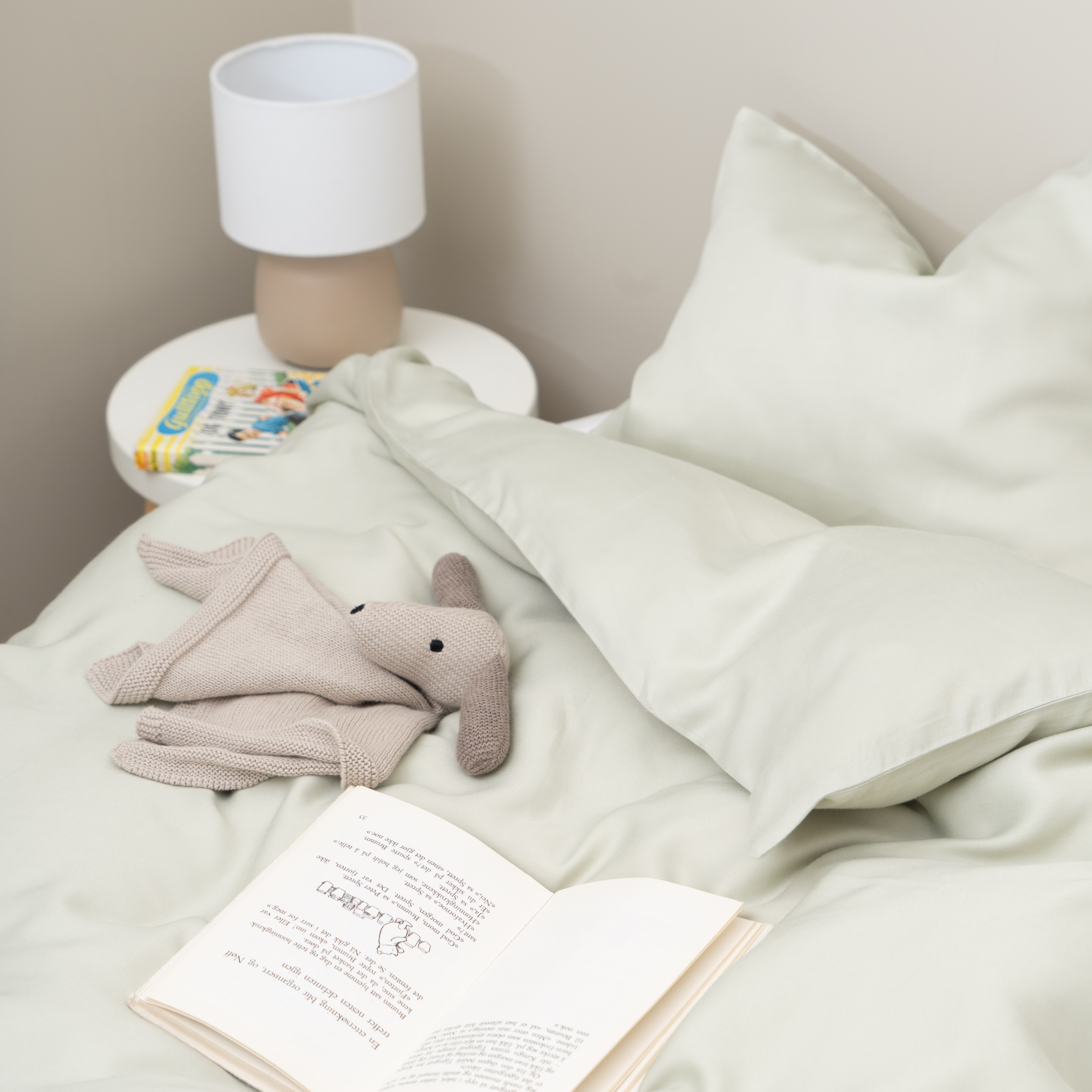 Lysegrønt sengetøysett med et lysebrunt strikket kosedyr og en åpen bok plassert på sengen.