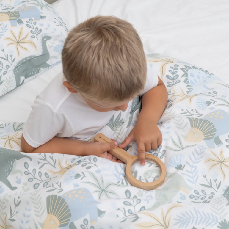 Mønstret sengetøy med blå og grønne dinosaurer, et barn i hvit t-skjorte holder et forstørrelsesglass i tre.