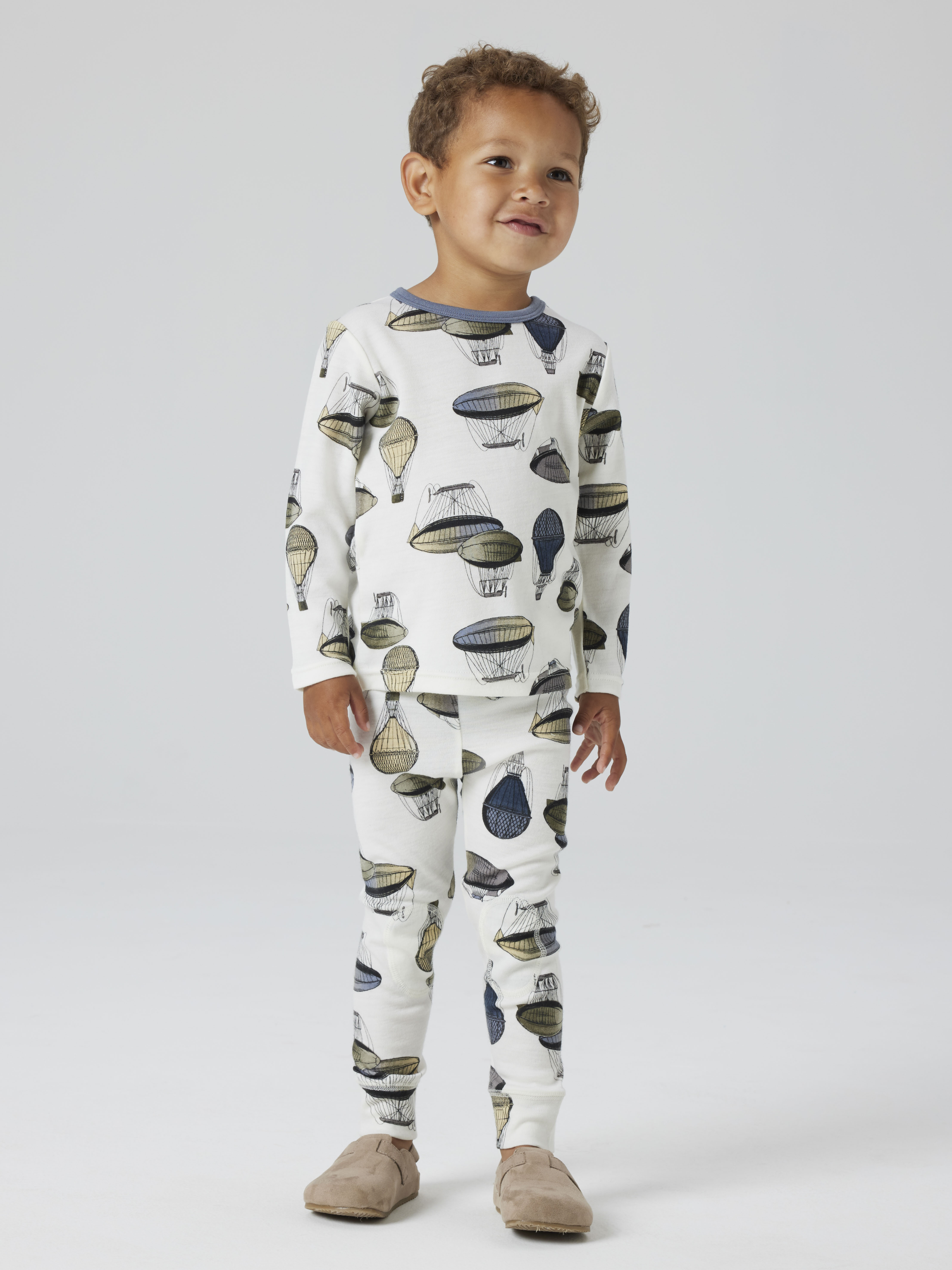 Barn har hvit todelt pyjamas med luftballongmønster, lange ermer, blå krage, og beige tøfler.