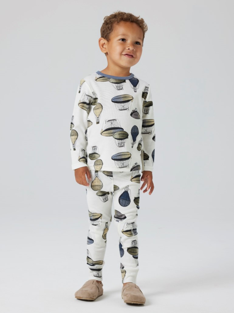 Barn har hvit todelt pyjamas med luftballongmønster, lange ermer, blå krage, og beige tøfler.