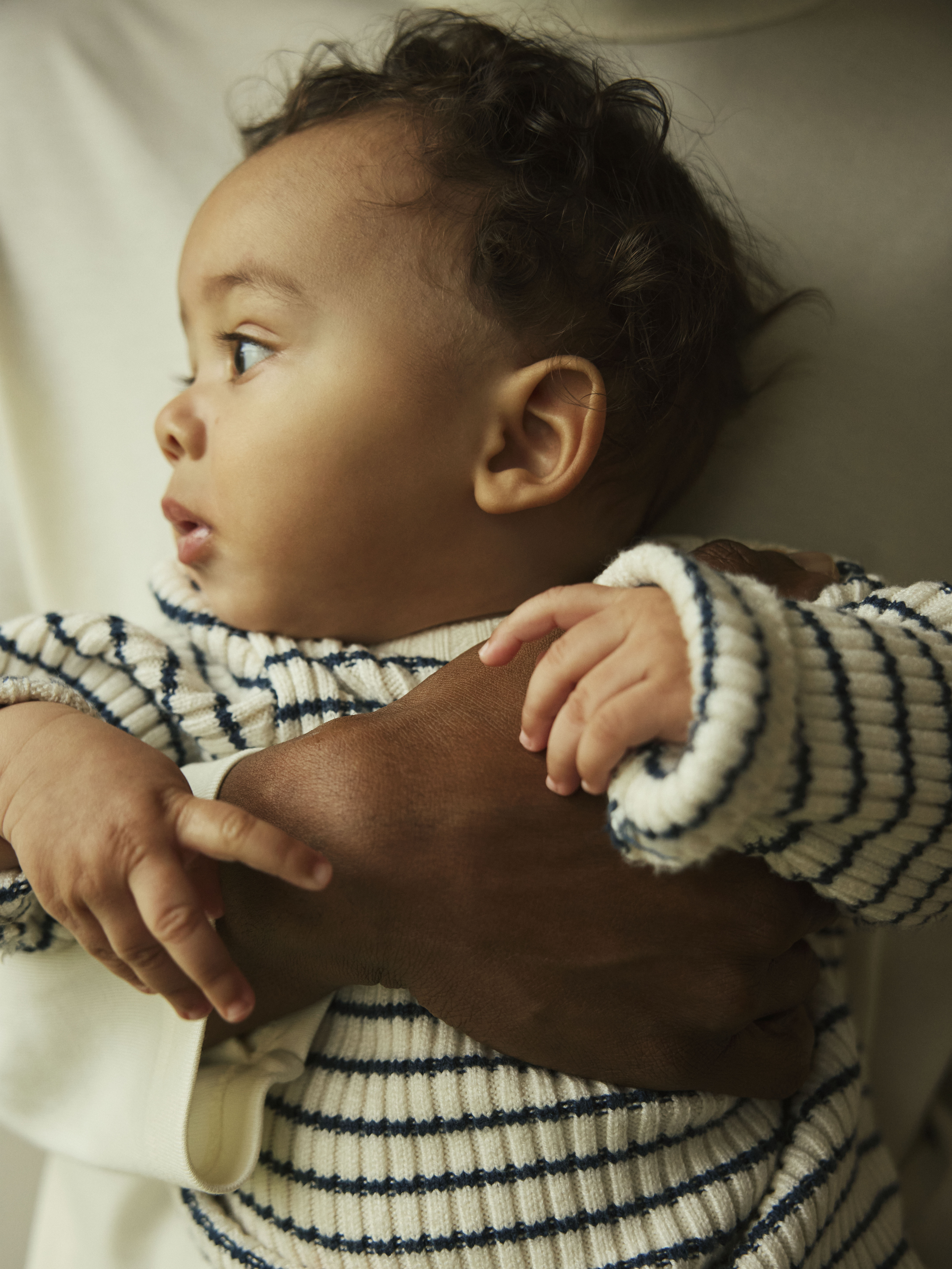 Baby har på seg en krem og marineblå stripete ribbestrikket genser, holdt av en voksen i kremfarget topp.