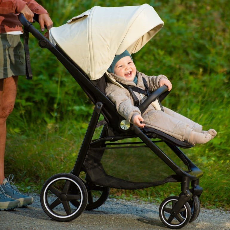Svart barnevogn med lys beige kalesje bærer en baby i beige vattert kjeledress, mørk turkis lue.