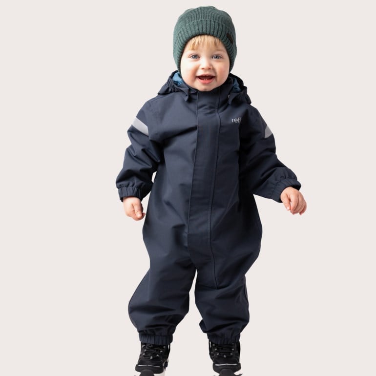 Forfra et barn i marineblå vanntett overall, en grønn strikket lue og svarte sneakers.