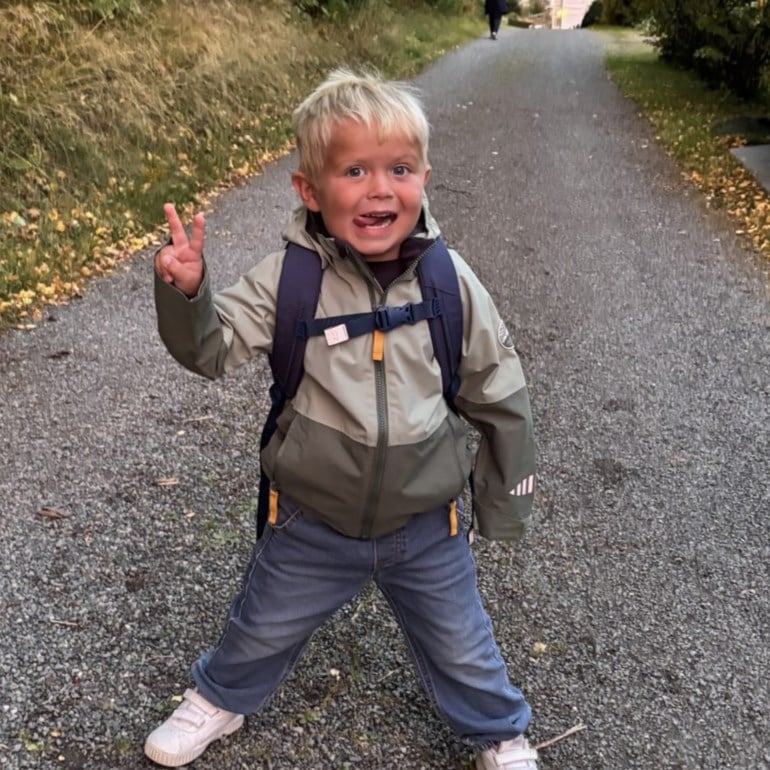 Barn bærer en tofarget olivengrønn utendørsjakke, en marineblå ryggsekk, blå jeans og hvite joggesko, forfra.