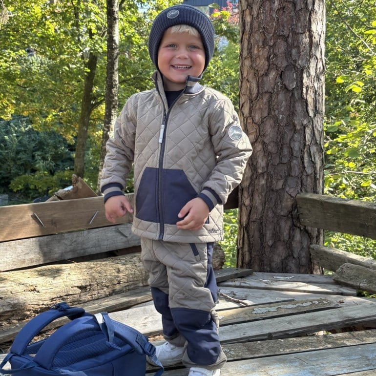 Barn i en vattert beige og marineblå jakke og matchende bukser, marineblå lue, og marineblå ryggsekk.