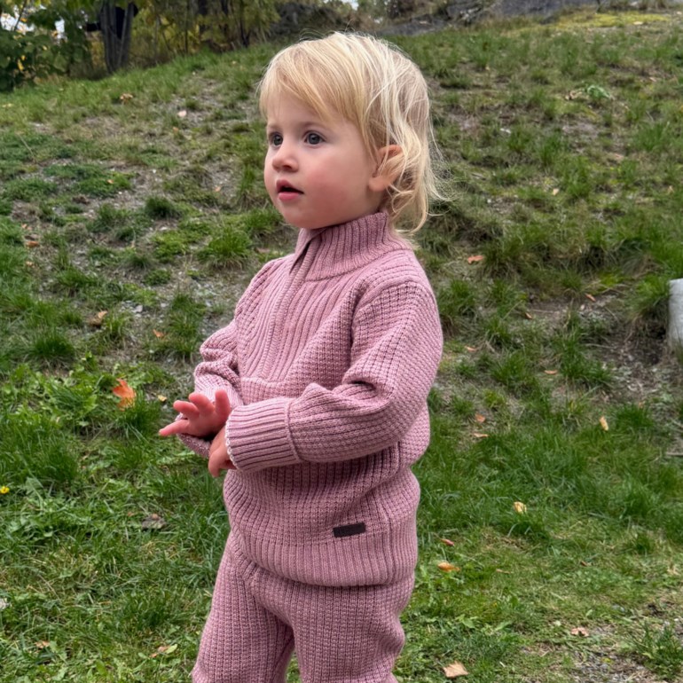 Barn bærer rosa strikket genser med halv glidelås, høy krage og matchende ribbestrikkede bukser.