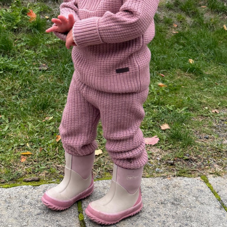 Barn i et støvrosa ribbestrikket sett med genser og bukser, samt tofargede beige og rosa gummistøvler.