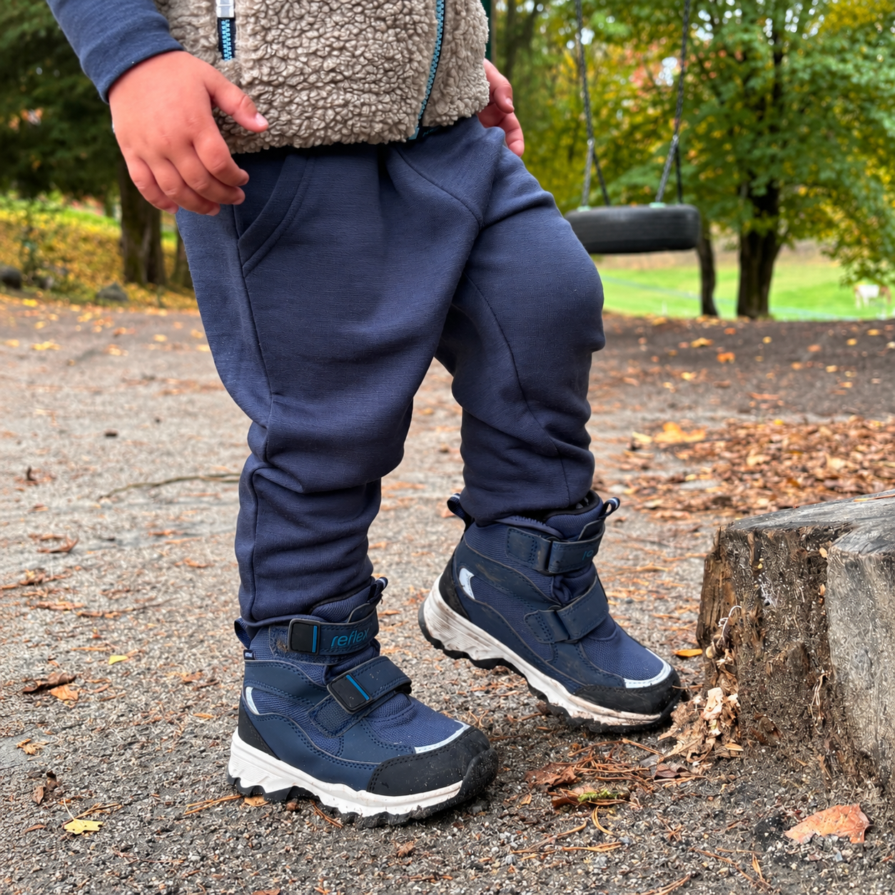 Barn har på seg mørkeblå vinterstøvler, mørkeblå joggebukse, langermet genser og en beige teddyvest.