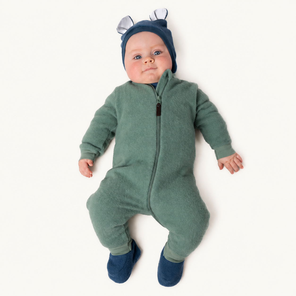 Baby i grønn ullfleece heldress med glidelås, mørkeblå lue med ører og mørkeblå tøfler.