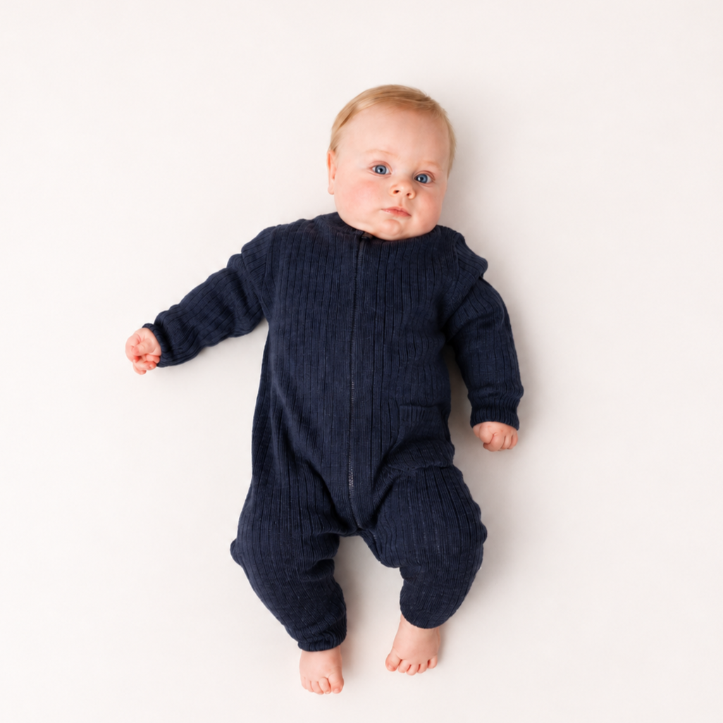 En baby har på seg en mørkeblå ribbet heldress med glidelås foran, liggende på hvit overflate.
