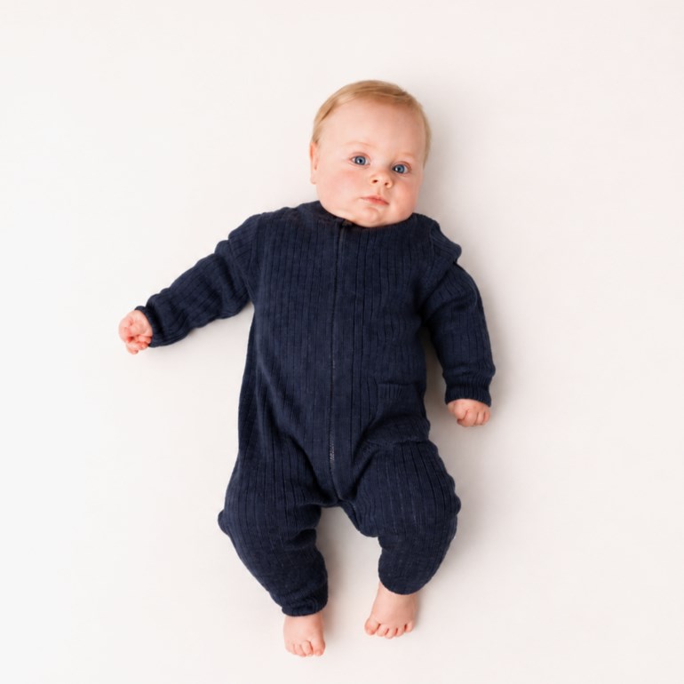 En baby har på seg en mørkeblå ribbet heldress med glidelås foran, liggende på hvit overflate.