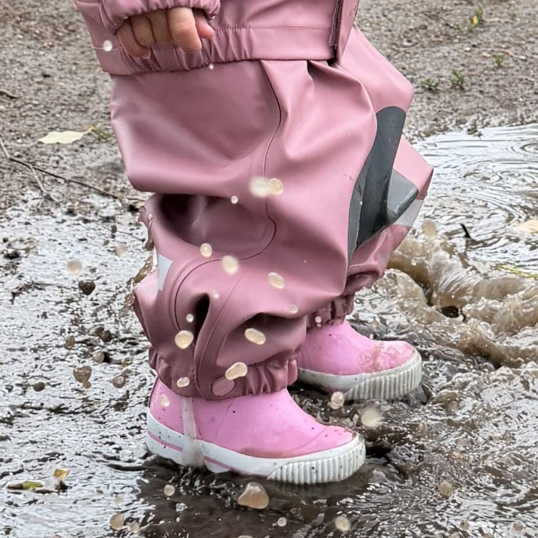 Barn i rosa vanntette regnbukser med mørke detaljer og rosa gummistøvler med hvite såler, plasker i sølepytt.