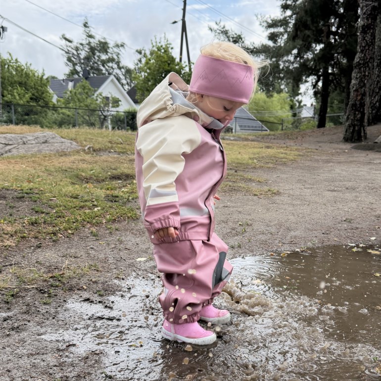 Barn har på seg rosa og beige vanntett overall med hette, rosa gummistøvler og rosa mønstret pannebånd, plaskende.