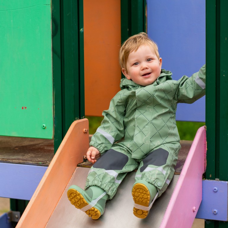 Baby har en grønnmønstret regndress med reflekser og mørke knelapper, pluss grønne støvler.