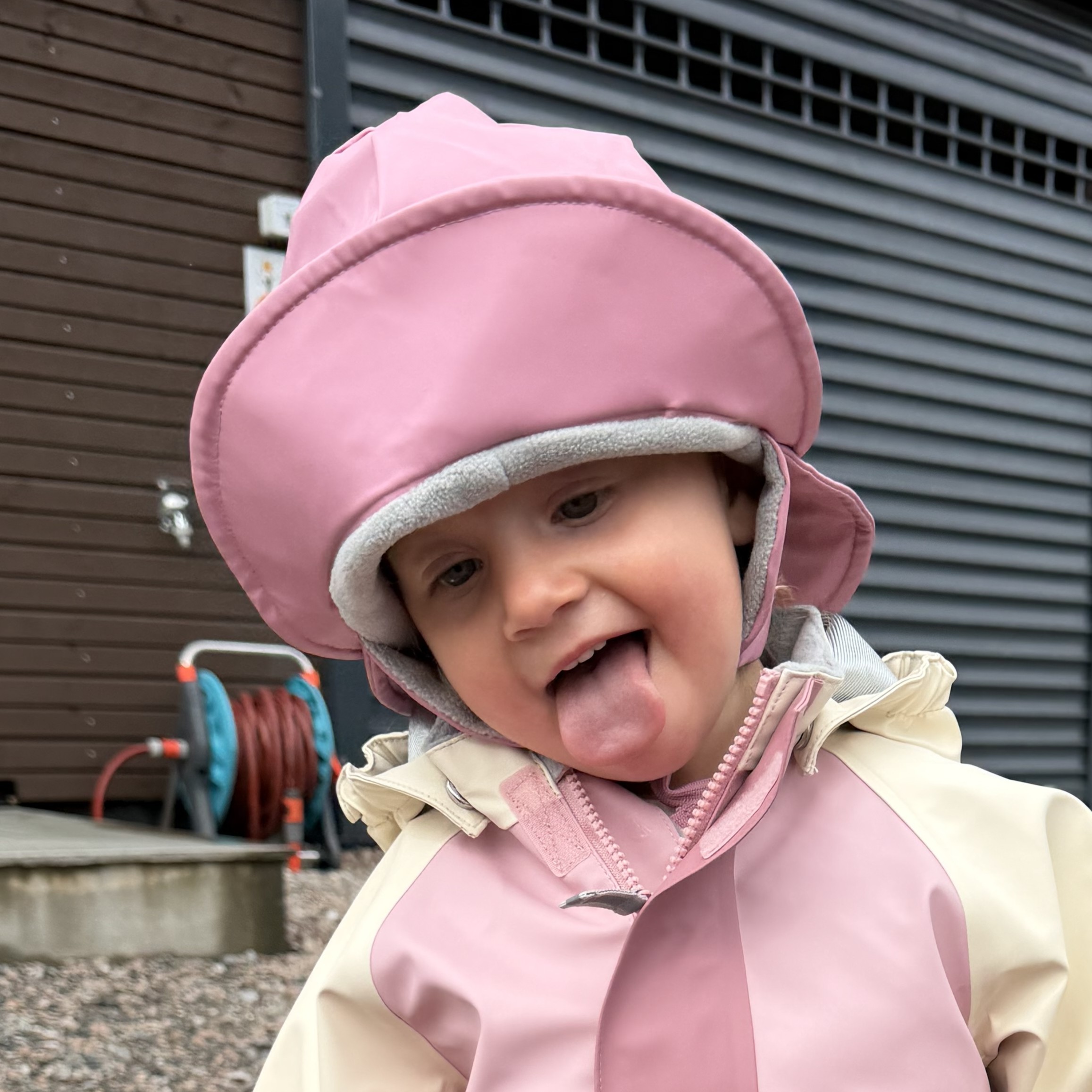 Barn med rosa regnhatt med grått fôr og rosa og beige regnjakke med glidelås foran.