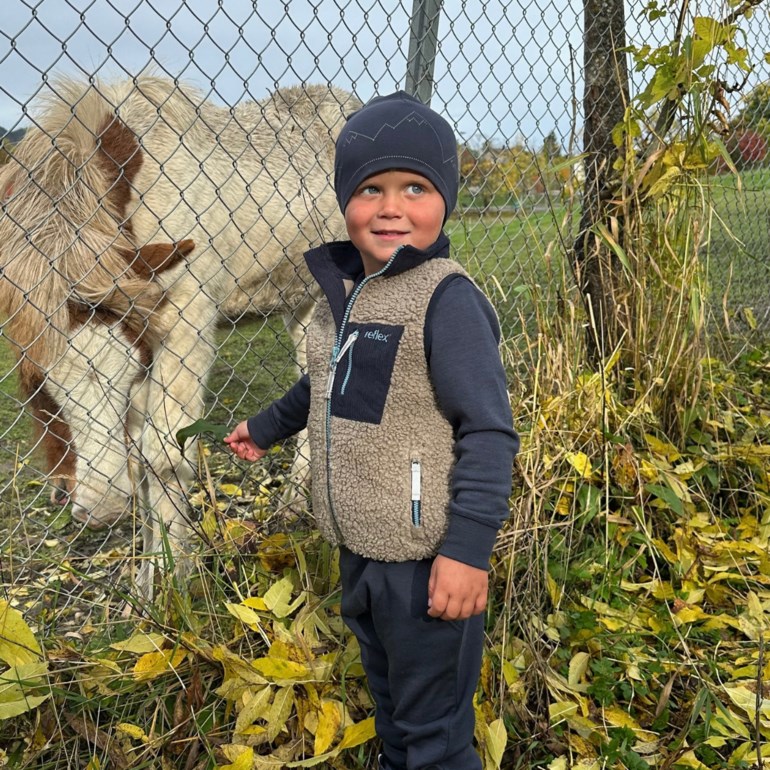 Barn i beige teddyvest med mørkeblå lomme, lyseblå glidelåser, mørkeblå genser, joggebukse og lue.